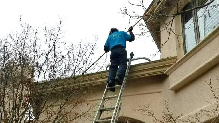 Technician on a ladder cleaning gutters along a home's roofline